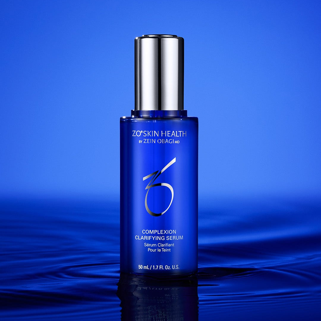 Zo Skin Health Complexion Clarifying Serum in a blue bottle with a silver cap, standing on a reflective dark blue surface against a blue gradient background.